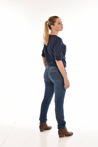 full length portrait of girl wearing simple blue shirt and jeans, standing in profile against a white studio background. - Zdjęcie, obraz