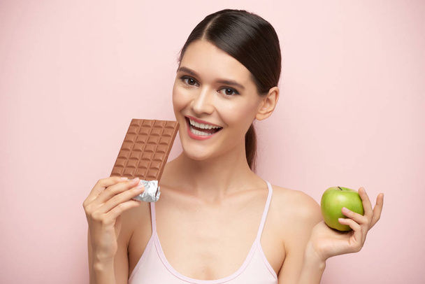 Gülümseyen genç kadın çikolata ve yeşil elma arasında seçme - Fotoğraf, Görsel