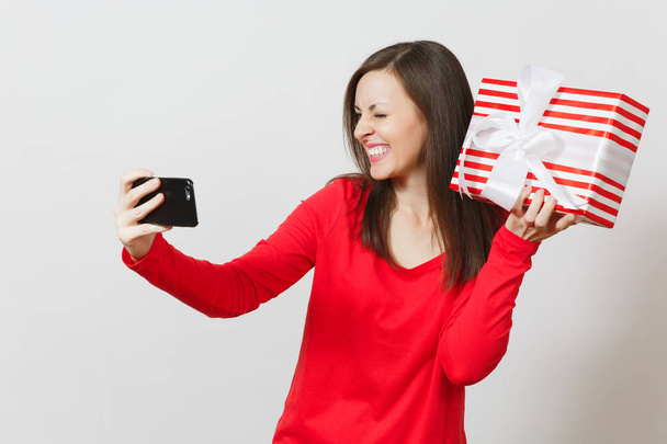 Woman doing selfie on mobile phone with red striped present box with gift isolated on white background. Copy space for advertisement. St. Valentine's Day, International Women's Day, holiday concept. - Photo, image