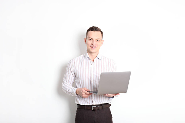 Smiling confident young man, no tie, holding grey laptop device and typing while standing against solid white wall. Wireless internet, wifi, enter password. Shaved, no facial hair. Isolated background - Fotó, kép