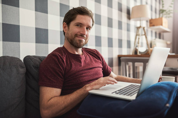 Smiling young man sitting alone on his living room sofa at home browsing the internet with laptop - Foto, Imagem