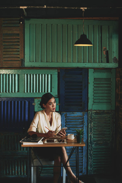 Bella donna d'affari vietnamita riflettere su nuove idee e prendere appunti quando si è seduti in un caffè atmosferico
 - Foto, immagini