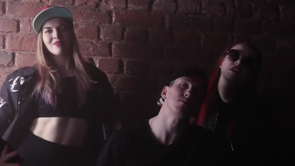 Young hip hop man in cap with face tattoos in front of brick wall with girls - Footage, Video