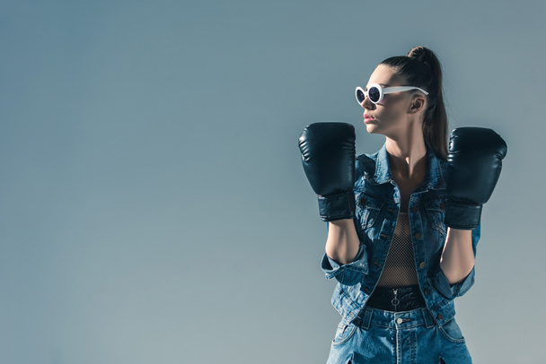 stylish girl in denim clothes and sunglasses posing with boxing gloves, isolated on grey - Foto, Bild