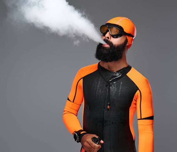 Retrato de um macho barbudo em terno de neoprene laranja e máscara de mergulho fumando um cachimbo eletrônico. Isolado sobre fundo cinzento
. - Foto, Imagem
