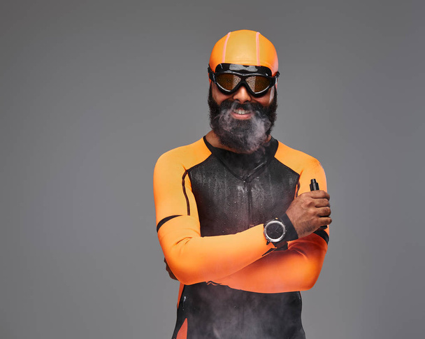 Porträt eines bärtigen Männchens in orangefarbenem Neopren-Taucheranzug und Tauchermaske, das eine elektronische Pfeife raucht. isoliert auf grauem Hintergrund. - Foto, Bild