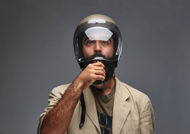 A bearded hipster in a motorcycle helmet smoking electronic cigarette. Isolated on a grey background. - Photo, Image