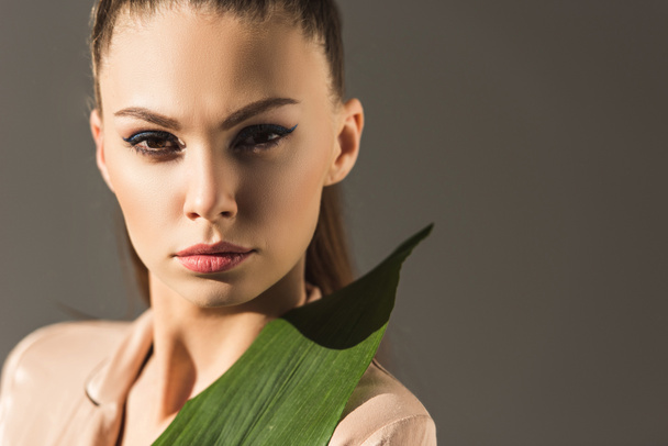 attractive girl with green leaf looking at camera, isolated on grey - Foto, afbeelding