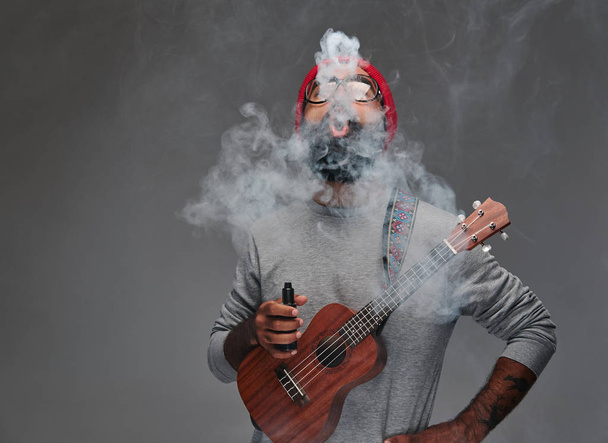 Retrato de um homem hipster barbudo em um chapéu vermelho e óculos de sol, jogando no ukulele. Isolado sobre fundo cinzento
. - Foto, Imagem