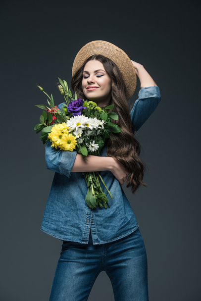 attractive smiling girl with closed eyes in denim clothes and straw hat holding bouquet of flowers, isolated on grey - Foto, imagen
