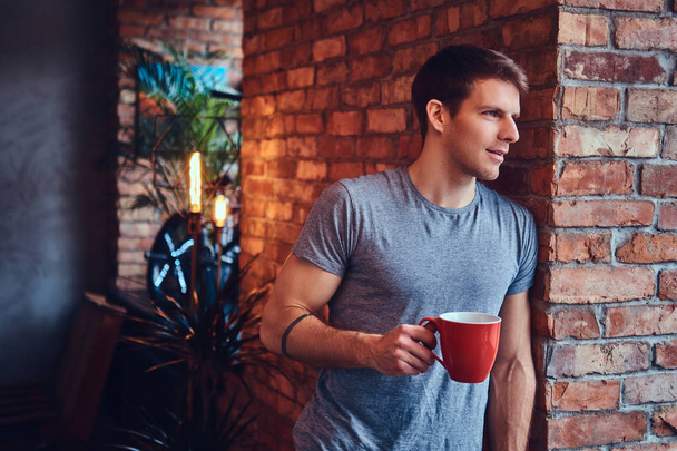 A stylish tattoed attractive man dressed in jeans and a t-shirt, leans against the brick wall, holding a cup of coffee. Smiling and looking away. - Foto, Imagen