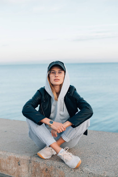 portrait of young woman in grey hoodie and sweatpants sitting on pier - Fotó, kép
