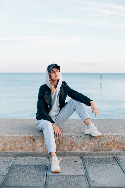 portrait of young woman in grey hoodie and sweatpants sitting on pier - Zdjęcie, obraz