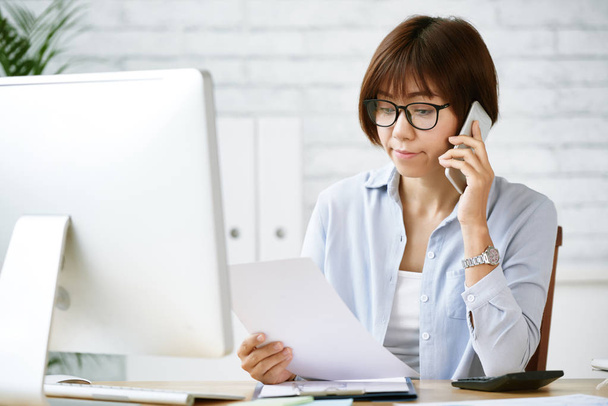 Портрет японской бизнес-леди в очках, разговаривающей по телефону и читающей документ
 - Фото, изображение