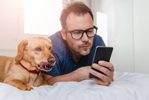 Man with glasses in blue shirt laying on the bed with the dog and using smart phone. - Foto, imagen