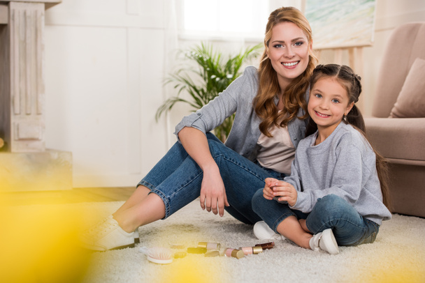selective focus of happy mother and daughter with cosmetics sitting on carpet and smiling at camera - Фото, зображення