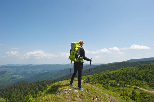 Conceito de estilo de vida ativo. Uma jovem mulher de preto com mochila verde brilhante e varas de trekking em pé no topo de uma colina, tendo a vista incrível das montanhas, floresta e céu
. - Foto, Imagem