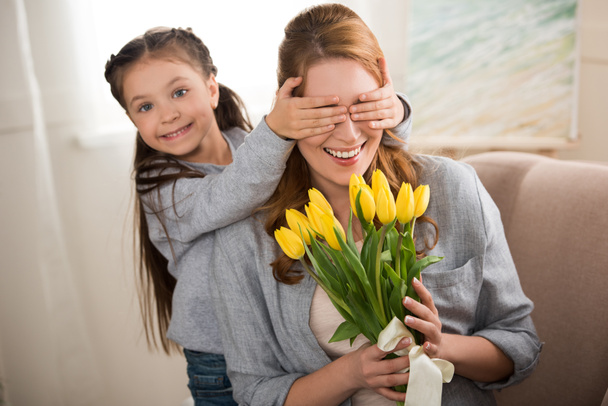 fröhliches Kind schließt Augen vor fröhlicher Mutter mit gelben Tulpen - Foto, Bild