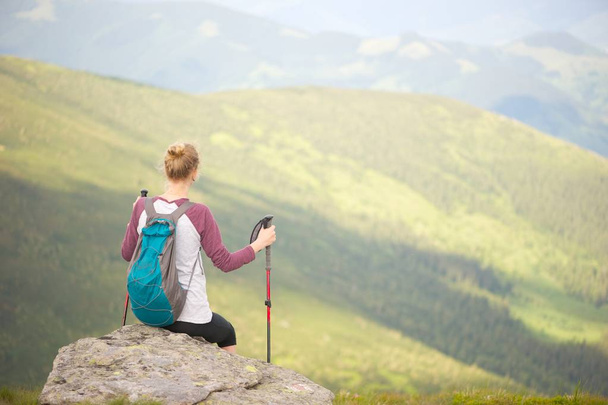 Uma jovem mulher sentada em uma rocha tendo a vista incrível das montanhas. Segurando paus de trekking; mochila em suas costas
. - Foto, Imagem
