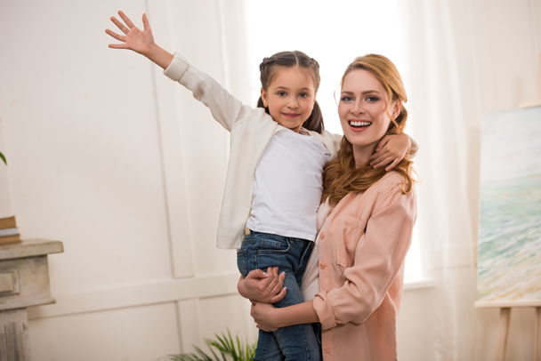 junge Frau trägt entzückende kleine Tochter und lächelt zu Hause in die Kamera - Foto, Bild