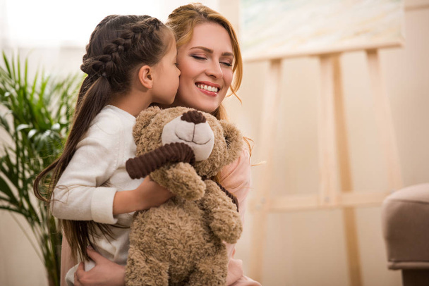 cute little child holding teddy bear and kissing happy mother at home - Foto, imagen