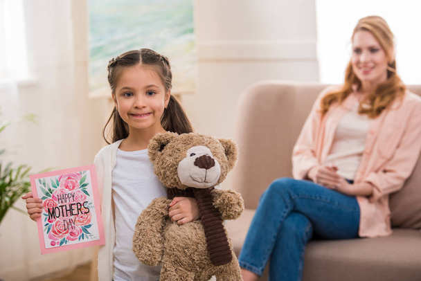 adorable child holding teddy bear and happy mothers day greeting card while mother sitting behind - Photo, Image