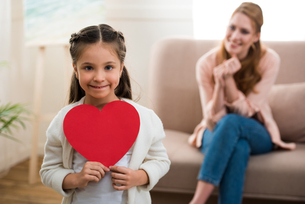 adorable child holding red heart symbol and smiling at camera while mother sitting behind - Photo, Image