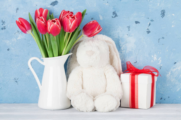 Red tulip flowers bouquet in pitcher, gift box and rabbit toy in front of stone wall.  Easter greeting card - Photo, Image