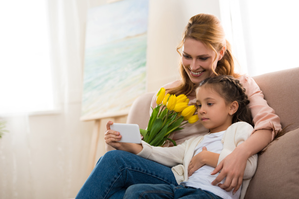Gelukkig moeder en dochter met gele tulpen met smartphone samen thuis - Foto, afbeelding