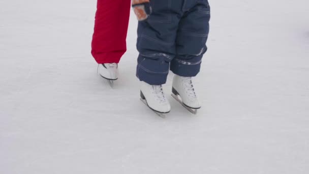 Los primeros pasos de un niño pequeño en patinaje sobre hielo. Mamá enseña patinaje y sostiene a su hijo en el hielo. Estilo de vida activo
 - Imágenes, Vídeo