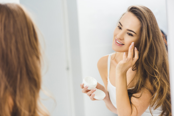 mirror reflection of beautiful smiling woman applying face cream - Photo, Image