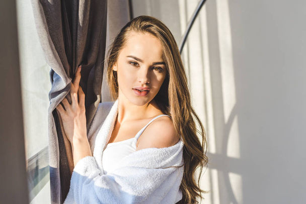 portrait of pretty young woman in bathrobe standing at window at home - Photo, Image