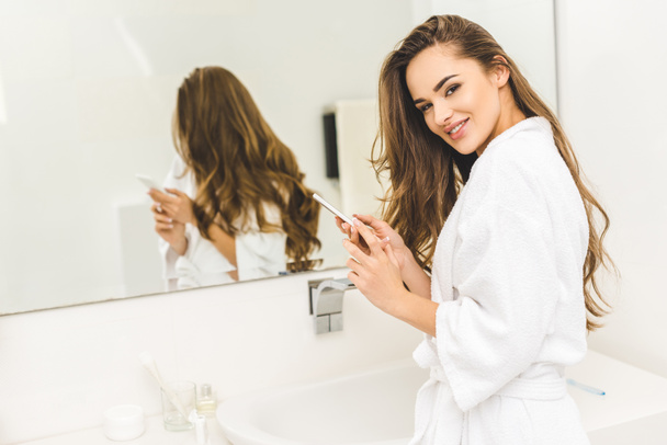 portrait of smiling woman in bathrobe with smartphone in hands in bathroom - Foto, Imagen