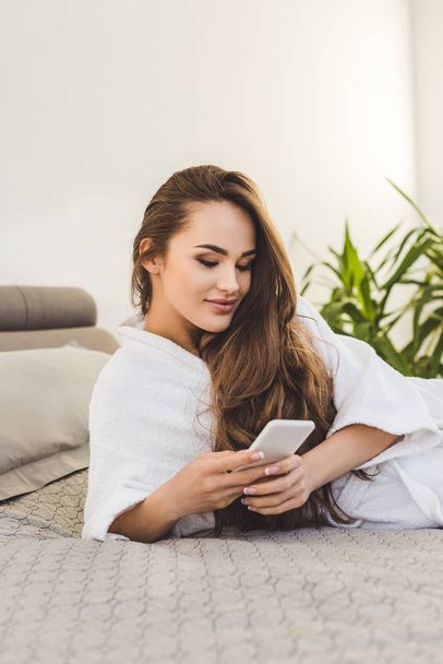smiling young woman in bathrobe with smartphone lying on bed - Fotografie, Obrázek