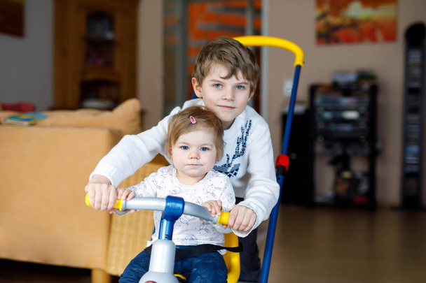 Μικρό παιδί αγόρι ώθηση bycicle ή τρίκυκλο με χαριτωμένο μωρό αδελφή. Μικρό παιδί κορίτσι και ο αδελφός παίζοντας μαζί στο σπίτι, σε εσωτερικούς χώρους. - Φωτογραφία, εικόνα