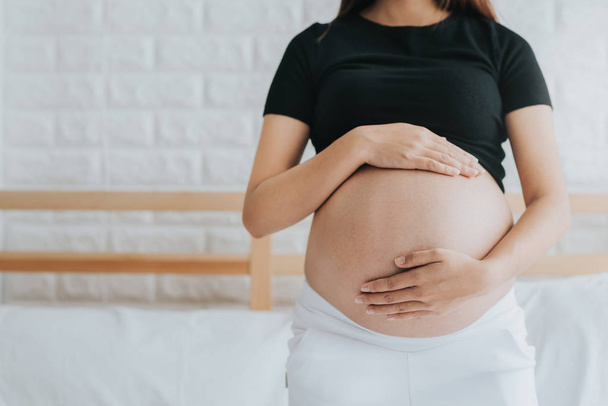 abgeschnittenes Bild einer schwangeren Frau, die ihren Schwangerschaftsbauch in der Nähe des Fensters zu Hause auf dem Bett hält - Foto, Bild