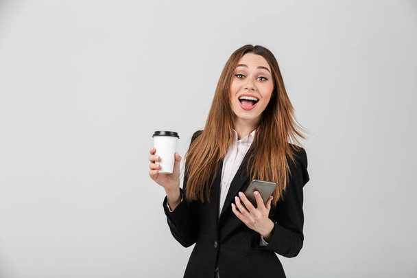Happy woman looking camera while holding cup of coffee and smartphone isolated - Zdjęcie, obraz