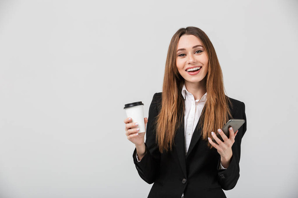 Smiling beautiful lady in business suit holding coffee and smartphone in hands isolated - Fotografie, Obrázek