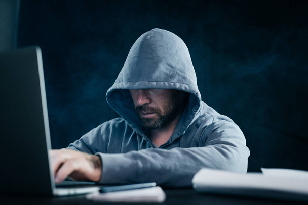 a man hacker in a sweatshirt with a hood sits behind a laptop in a dark room - Foto, afbeelding