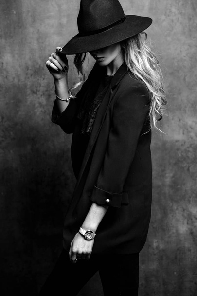 Beautiful young mysterious blond girl in black hat and black jacket on gray background. Eyes are covered with a hat. Manicure - long red nails, nail polish. Fashion, beauty. - Valokuva, kuva