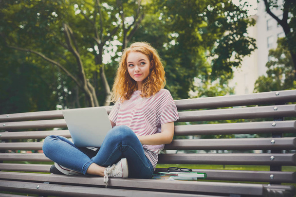 Молодая рыжая девушка с ноутбуком сидит на скамейке в парке на открытом воздухе
 - Фото, изображение
