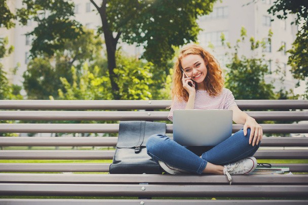 Kızıl saçlı çekici kız açık havada parkta bankta dizüstü bilgisayar ile telefon oturma üstünde konuşuyor - Fotoğraf, Görsel