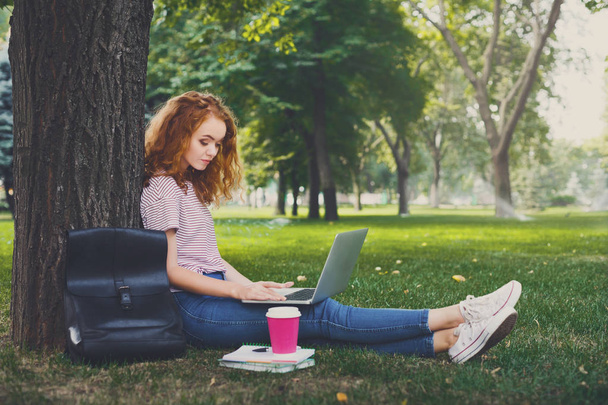 Jovem estudante ruiva trabalhando sentado debaixo de uma árvore
 - Foto, Imagem