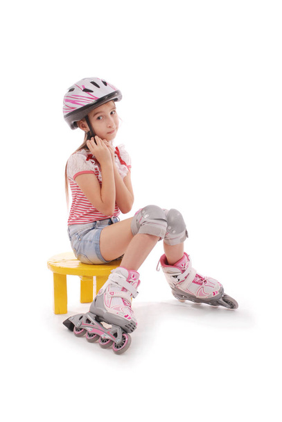Charming broun-haired girl of school age in short jeans shorts and a pink t-shirt sitting on the chair and tries to foot roller skates. - Photo, Image