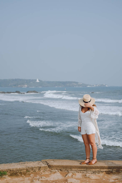 Girl in a hat waving in the wind light white clothes walking around the ocean - 写真・画像