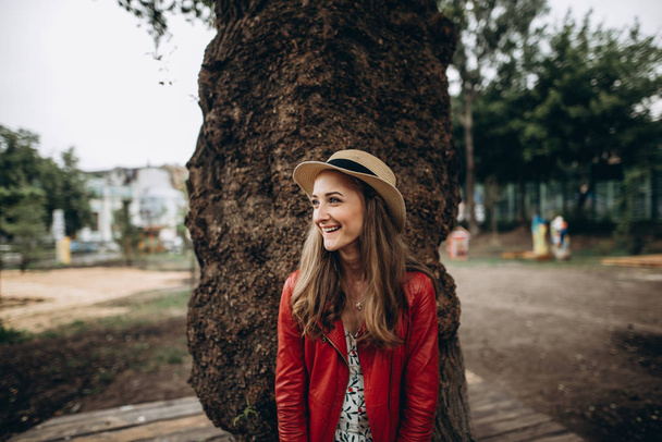 Close-up photo of a cute smiling blonde girl in a red jacket, with a hat on her head, stands near a big tree and looks aside - Zdjęcie, obraz