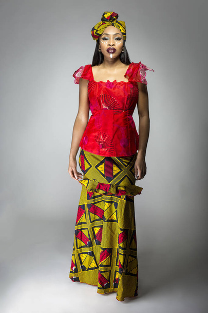 Black female showing african pride by wearing a traditional Nigerian dress and head scarf with tribal face markings or cosmetic makeup.  The costume is red and yellow and shows cultural fashion. - 写真・画像