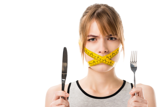 mujer joven con cinta métrica atada alrededor de la boca sosteniendo tenedor y cuchillo aislado en blanco
 - Foto, imagen