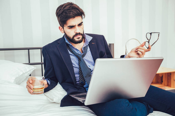 Werken in een hotelkamer. Zeker jonge zakenman in pak en stropdas bezig met laptop zittend op het bed in hotelkamer - Foto, afbeelding