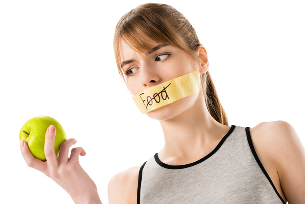 mladá žena s stick páska s přeškrtnutý slovo jídlo pokrývající ústa v ruce při pohledu na apple izolované na bílém - Fotografie, Obrázek